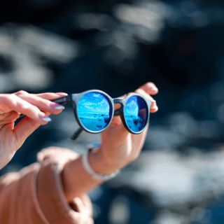 Some hands holding up sunglasses facing the sea. You can see the sea refleted in the coating of the sunglasses